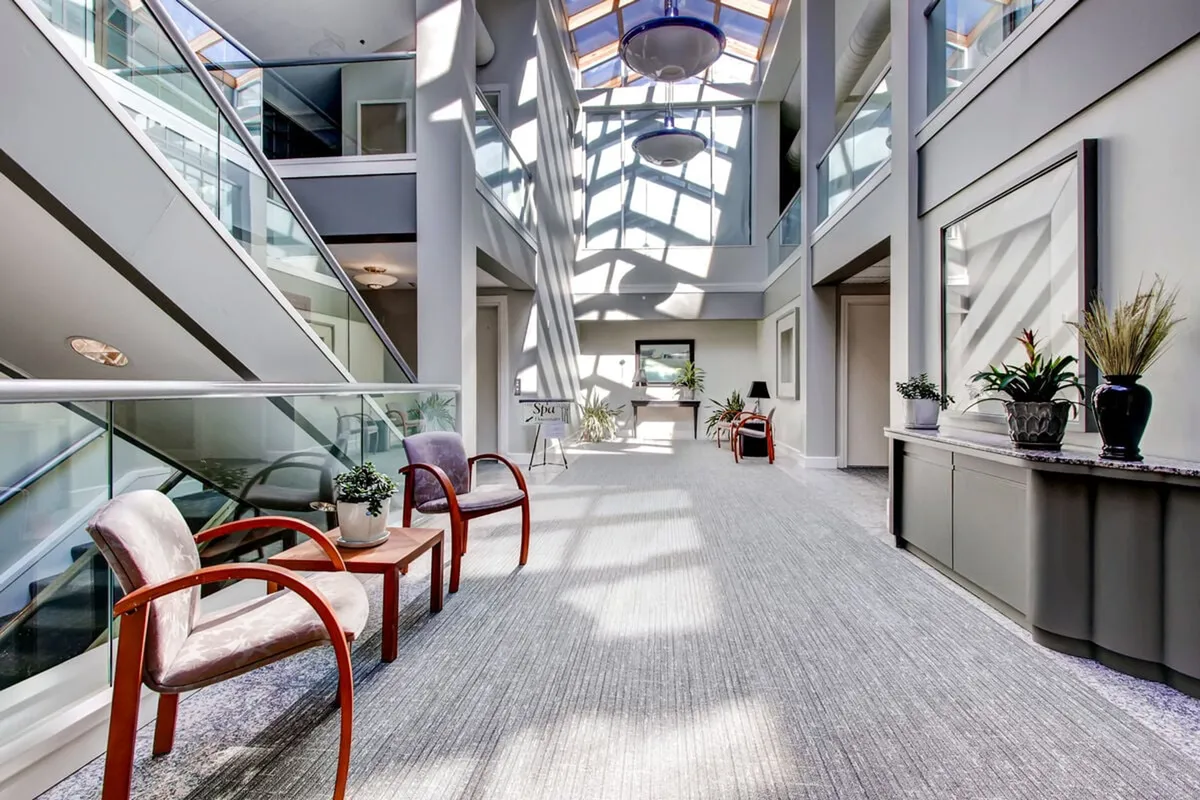 A tranquil, light-filled interior lounge area with skylights, lush indoor plants, comfortable chairs, and a decorative console table. The calm, welcoming mood suggests a space ideal for unwinding after a spa treatment or relaxing in between fitness activities.