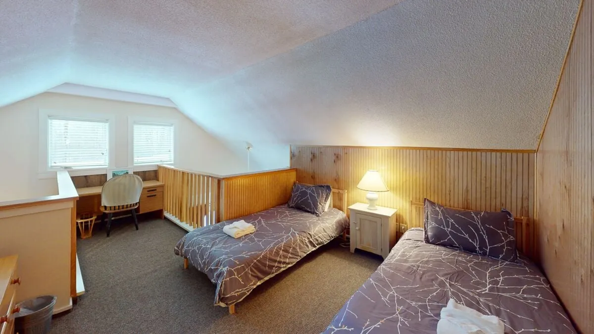 A tranquil bedroom with two neatly made twin beds, a central nightstand with a lamp, warm wood paneling, and a window area that brings in soft daylight. The room feels both inviting and flexible, ideal for families or groups.