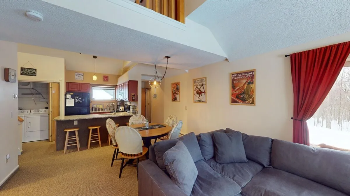 A wide, inviting open-plan space featuring a large sectional sofa, a dining table with chairs, and a visible kitchen area with bar stools. Large windows with red curtains bring in natural light and offer glimpses of the snowy outdoors. Decorative wall posters add a personal, homey touch. The scene hints at family gatherings, cozy meals, and relaxation after a day outside.