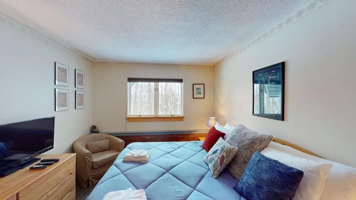 A cozy bedroom with a bed sporting a blue quilt, positioned near a large window overlooking the lush Vermont woods. A TV unit sits nearby, and the overall space is designed for relaxation and quiet moments, with abundant natural light and a tranquil forest view.