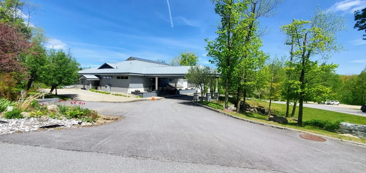 This exterior image showcases the modern architectural design of the resort building, framed by lush greenery and a curved driveway leading toward the entrance. The inviting landscaping and serene atmosphere suggest a welcoming arrival experience.