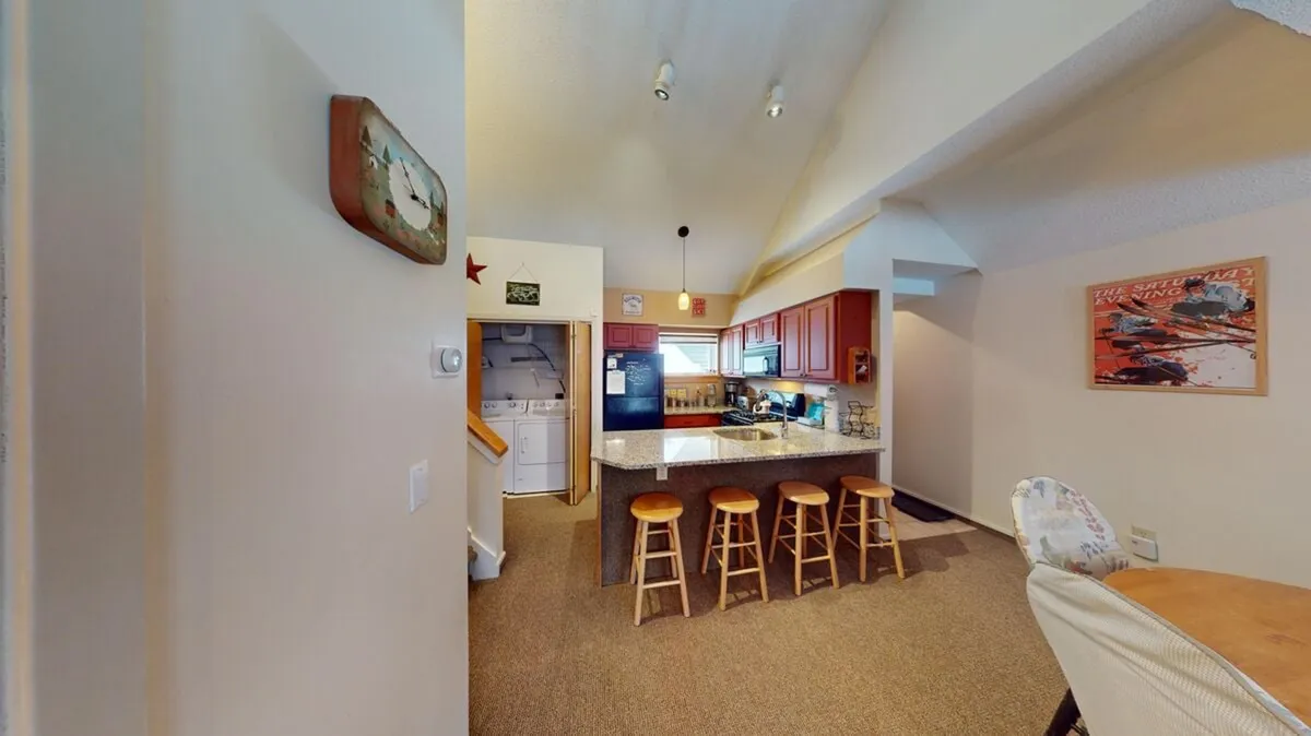 A bright and warm kitchen scene featuring the kitchen island with stools, black appliances, a window, and the adjoining dining area. The space is arranged for socializing, family meals, and seamless movement between kitchen and dining.
