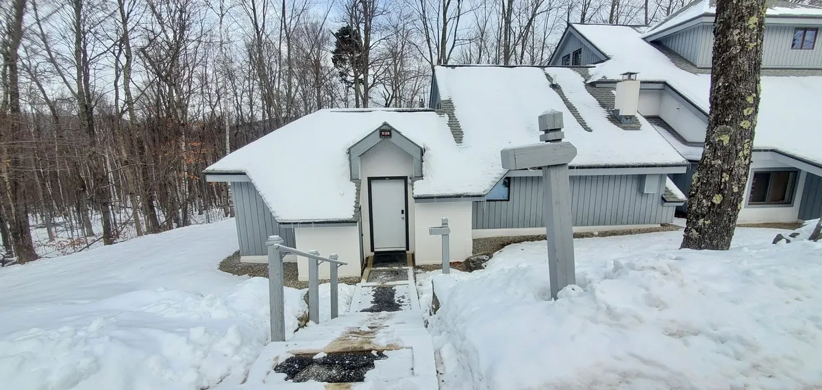 A picturesque exterior view of the property, featuring a snow-covered roof, entrance door, and wooden steps. The scene is blanketed in fresh snow, evoking the charm of a classic Vermont winter getaway. The inviting look of the home suggests a cozy retreat after a day on the slopes.
