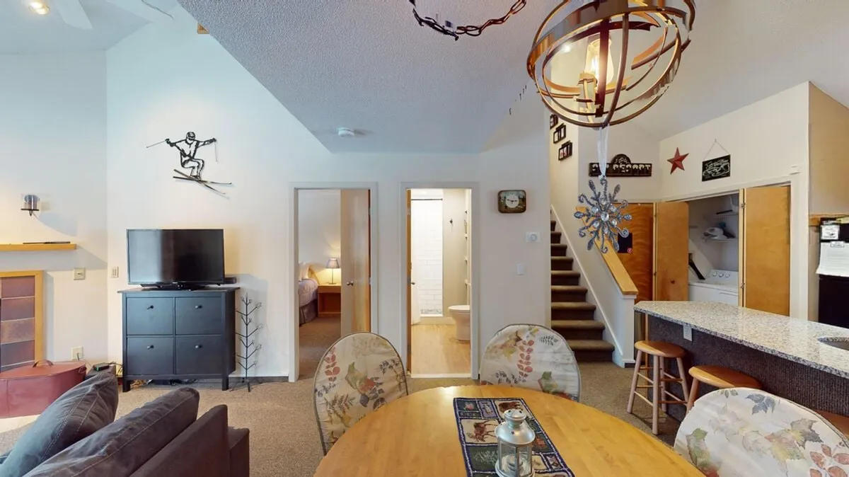 A welcoming living area featuring a comfortable sofa, a dining table ideal for shared meals or board games, a television for movie nights, and a staircase suggesting access to multiple levels. The space glows with warm lighting and hints at an inviting, communal atmosphere.