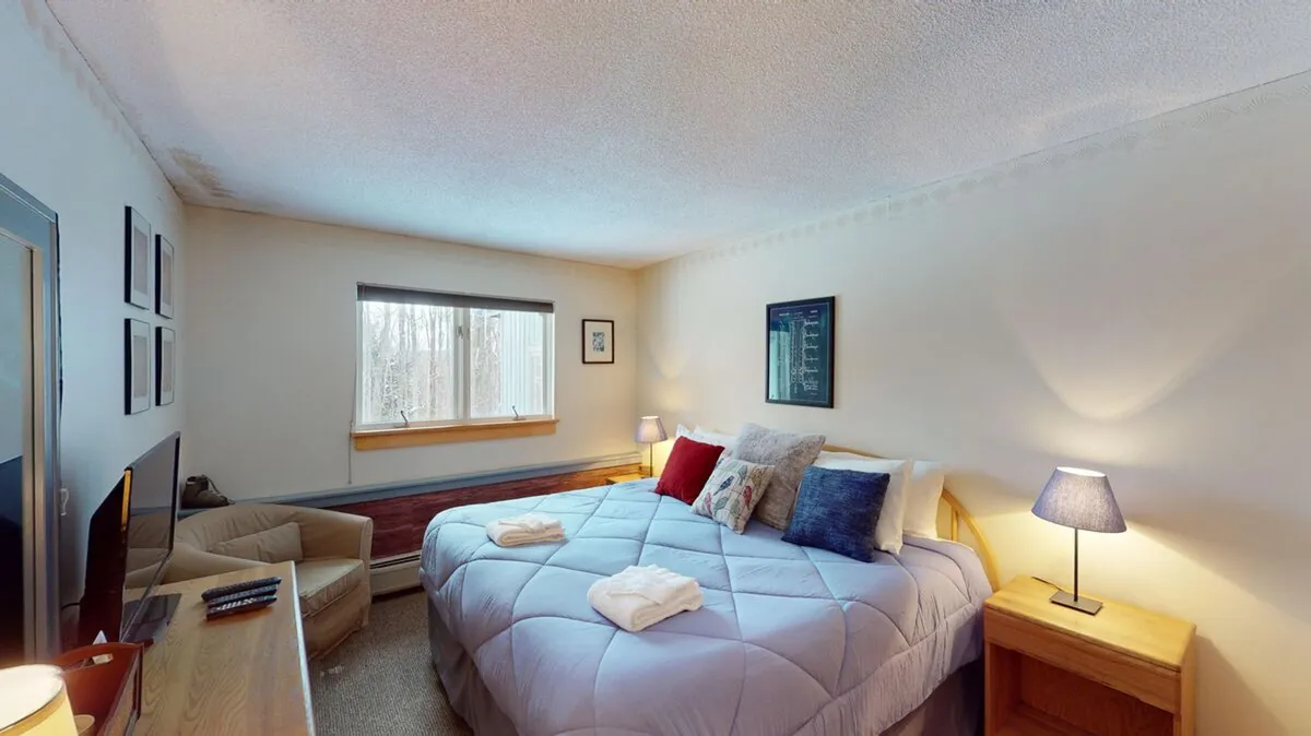 A serene bedroom featuring a comfortable bed with crisp linens, a window letting in natural light, inviting artwork, a lamp for ambient lighting, and a cozy chair creating a nook for reading or relaxing. The space exudes tranquility and privacy, perfect for unwinding after a day outdoors.